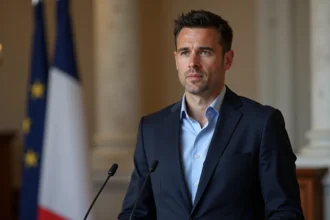 Portrait de Sébastien Delogu en veste marine devant un décor institutionnel français, podium et drapeaux tricolores en arrière-plan